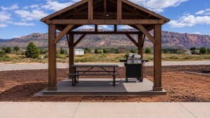Ein hölzerner Pavillon mit Picknicktisch und Grill, vor einer gebirgigen Kulisse.