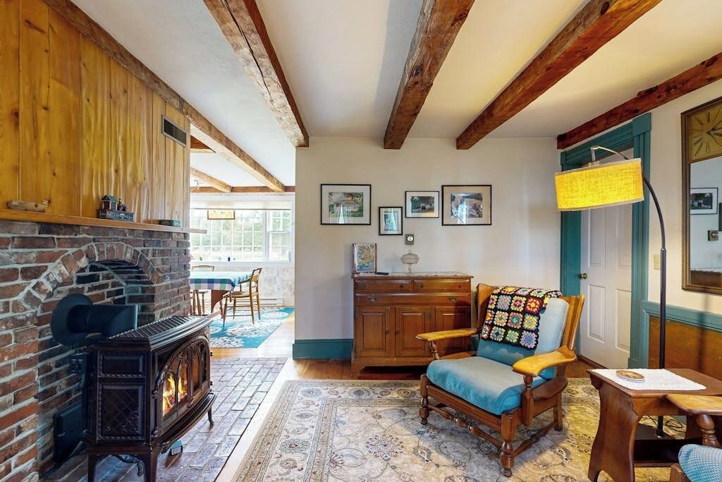 A cozy room with a fireplace, wooden beams, a rocking chair, and a dining area.