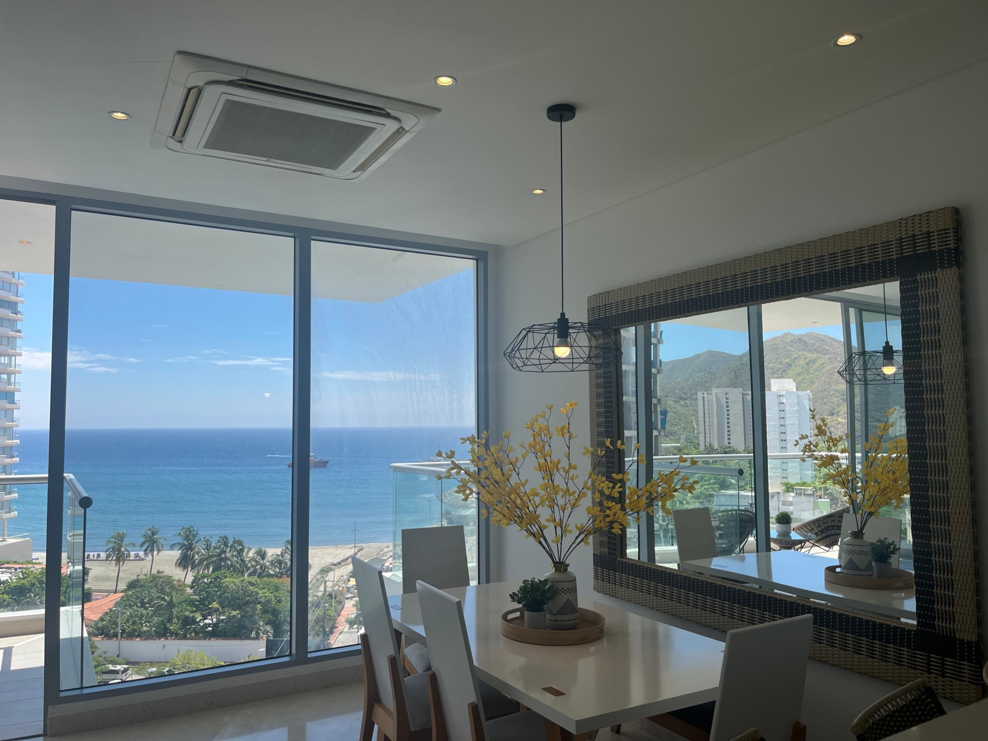 A modern dining area with a large window overlooking the ocean, a dining table, chairs, a vase with yellow flowers, and a hanging light fixture.