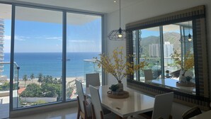Ein moderner Essbereich mit einem großen Fenster zum Meer, einem Esstisch, Stühlen, einer Vase mit gelben Blumen und einer Pendelleuchte.