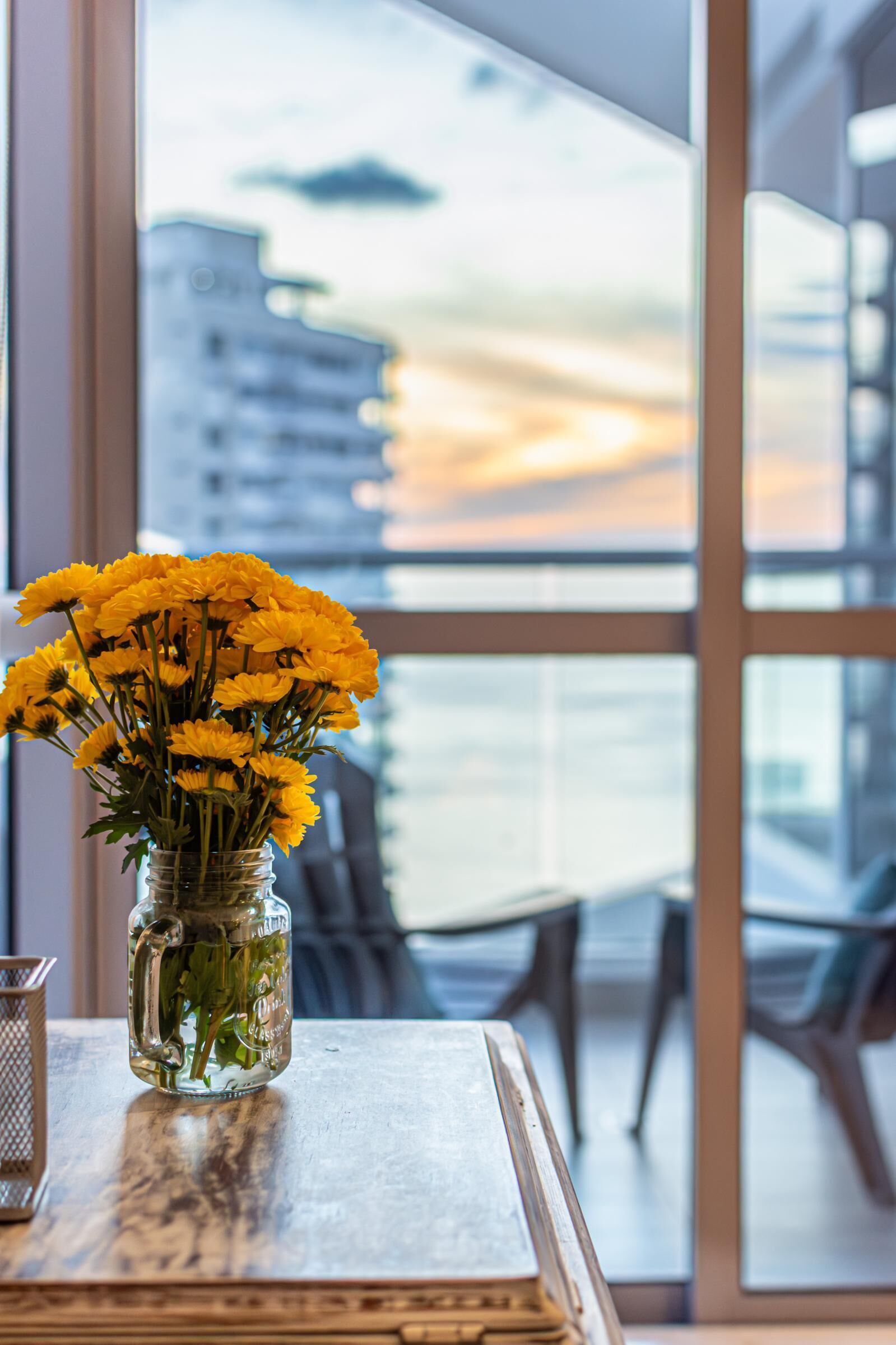 A clear view of a cityscape through large windows, a vase with yellow flowers on a table, and a metal candle holder.
