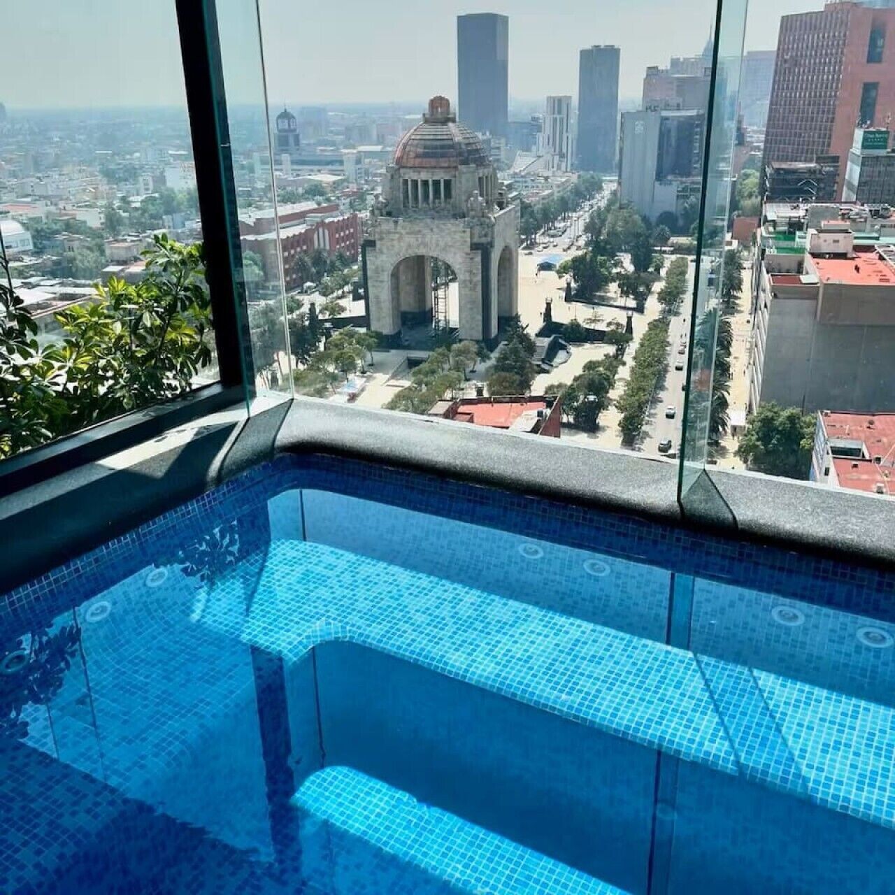 Une vue depuis une fenêtre d’un immeuble de grande hauteur, donnant sur un paysage urbain avec une imposante arche historique, une piscine et des bâtiments modernes.