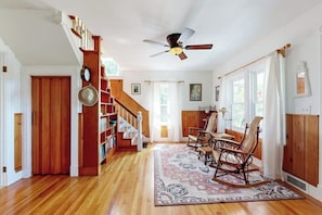 A spacious living room with a wooden staircase, a ceiling fan, rocking chairs, and a patterned rug. - Coastal Serenity Retreat with Ocean Views, Spacious Porch and Modern Comforts (Sullivan)