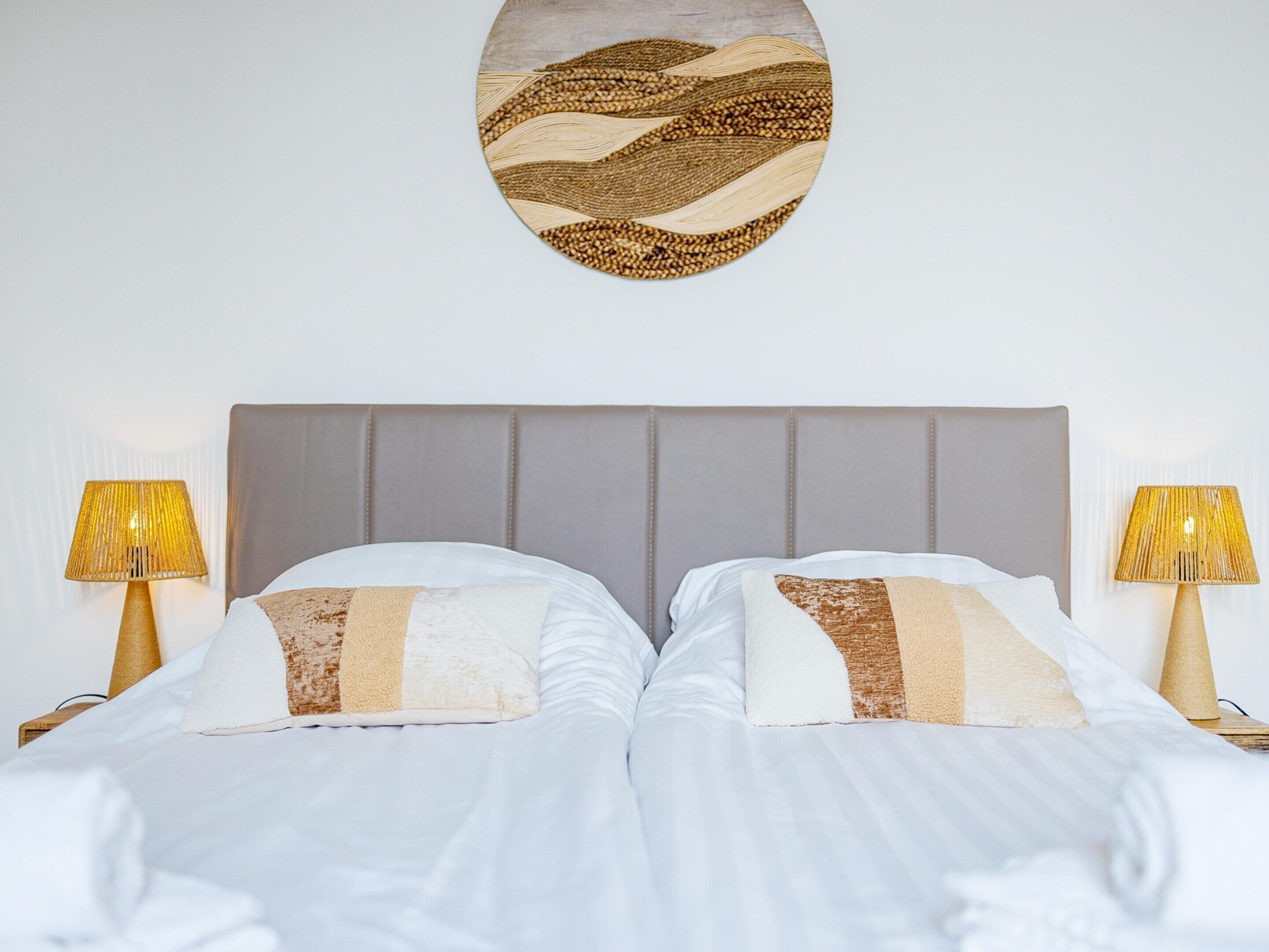 A bed with a grey headboard, two bedside lamps, and a wall-mounted artwork.
