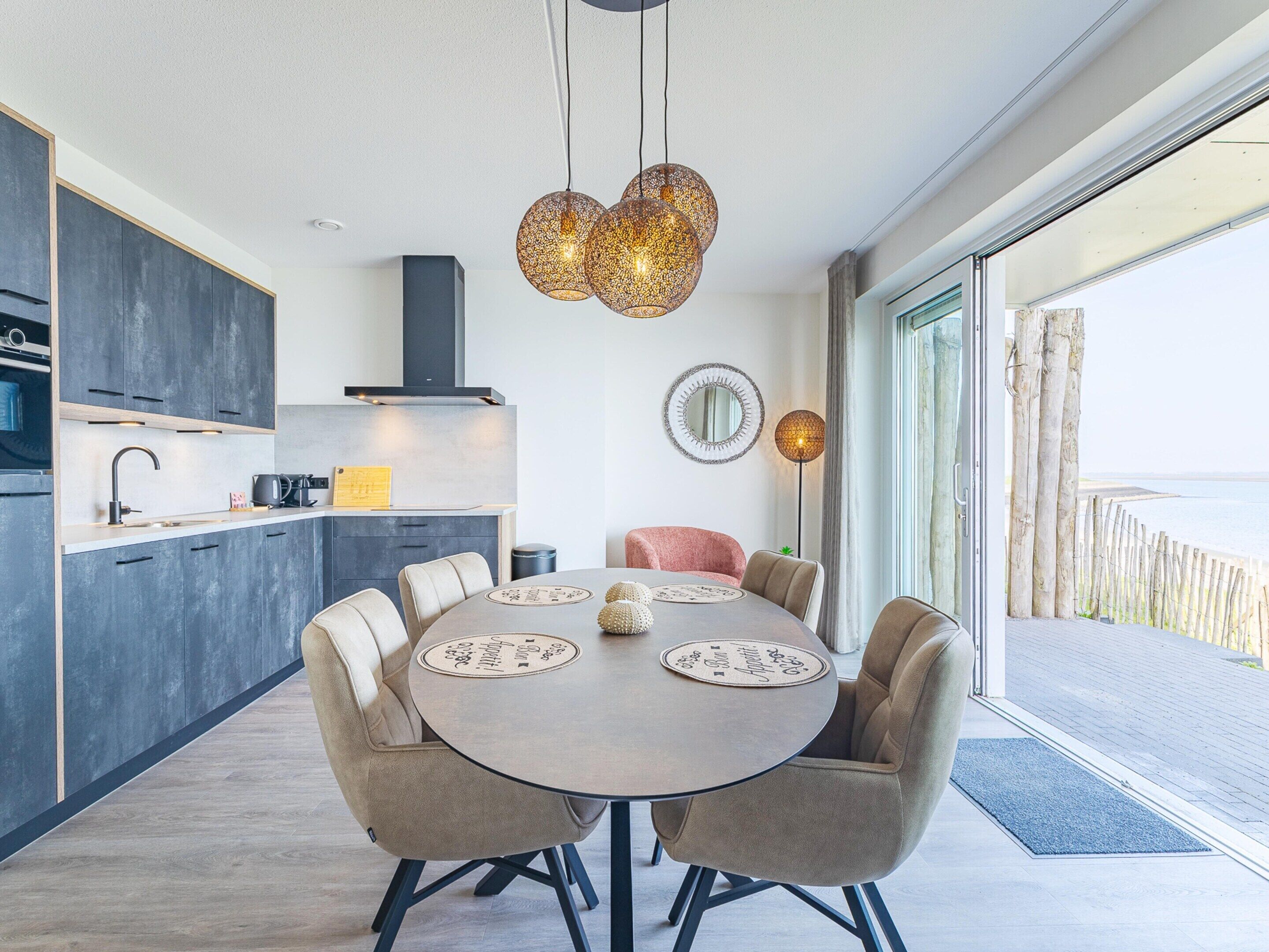 A modern kitchen with dark cabinets, a round dining table with chairs, and a view of the sea through large windows.