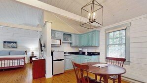 A room with a bed, a dining table with chairs, a kitchen area with light blue cabinets, and a ceiling light fixture. - Charming dog-friendly studio cabin with firepit and outdoor activities (Northport)