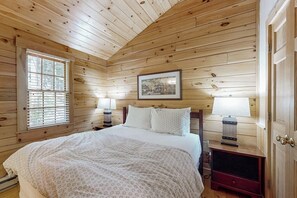 Ein Schlafzimmer mit Holzwänden, einem Bett, zwei Nachttischlampen, einem gerahmten Bild an der Wand und einem Fenster mit Jalousien.