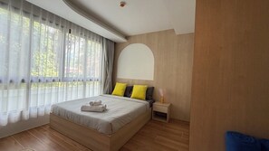 A modern bedroom with a large bed, a nightstand, a wall-mounted arch, and a window with sheer curtains. - Kamala Beach Residence A202 (Tambon Kamala)