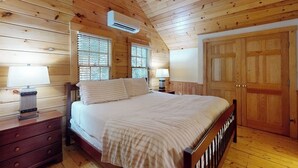 Un dormitorio de cabaña de madera con una cama, dos mesitas de noche, un aparador y un equipo de aire acondicionado, además de una puerta de madera grande.