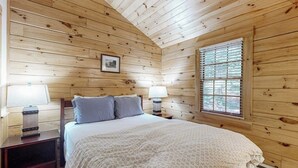Un dormitorio de cabaña de madera con una cama, dos mesitas de noche, una ventana con persianas y un cuadro enmarcado en la pared.