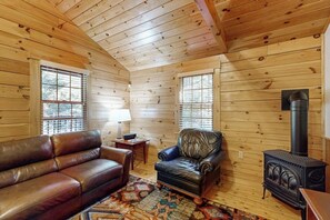 A log cabin interior with a leather sofa, armchair, and a wood-burning stove.