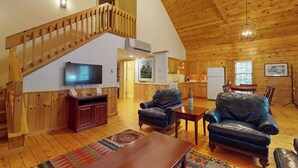 A spacious living area with a wooden staircase, a television, leather sofas, a dining table, and a kitchen in the background. - Bobcat Cabin, dog-friendly cabin retreat with full kitchen, outdoor activities (Northport)