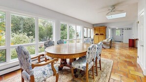 Uma área de refeições com uma mesa redonda de madeira e cadeiras estampadas, janelas amplas com vista para o verde e um ventilador de teto.