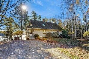 A single-story house with a sloped roof, a front porch, and a gravel driveway. - Charming riverfront home with cozy interiors, dock, and scenic deck views (Westport Island)