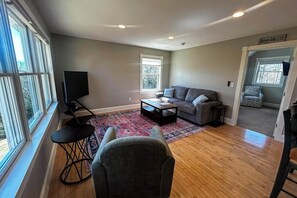 A living room with a sofa, coffee table, TV, and a view of the outdoors. - Charming coastal cottage with deck, grill, firepit, and jetted tub (Trevett)
