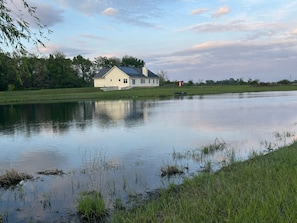 Una casa frente a un lago tranquilo, con césped al frente y árboles en el fondo.