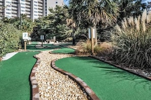 Un campo de mini golf con césped artificial verde, un camino de grava y una palmera.