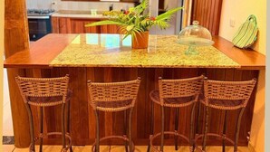 A kitchen with wooden cabinets, a granite countertop, a potted plant, a glass display case, and wicker bar stools. - Beautiful Leisure House (Trancoso)