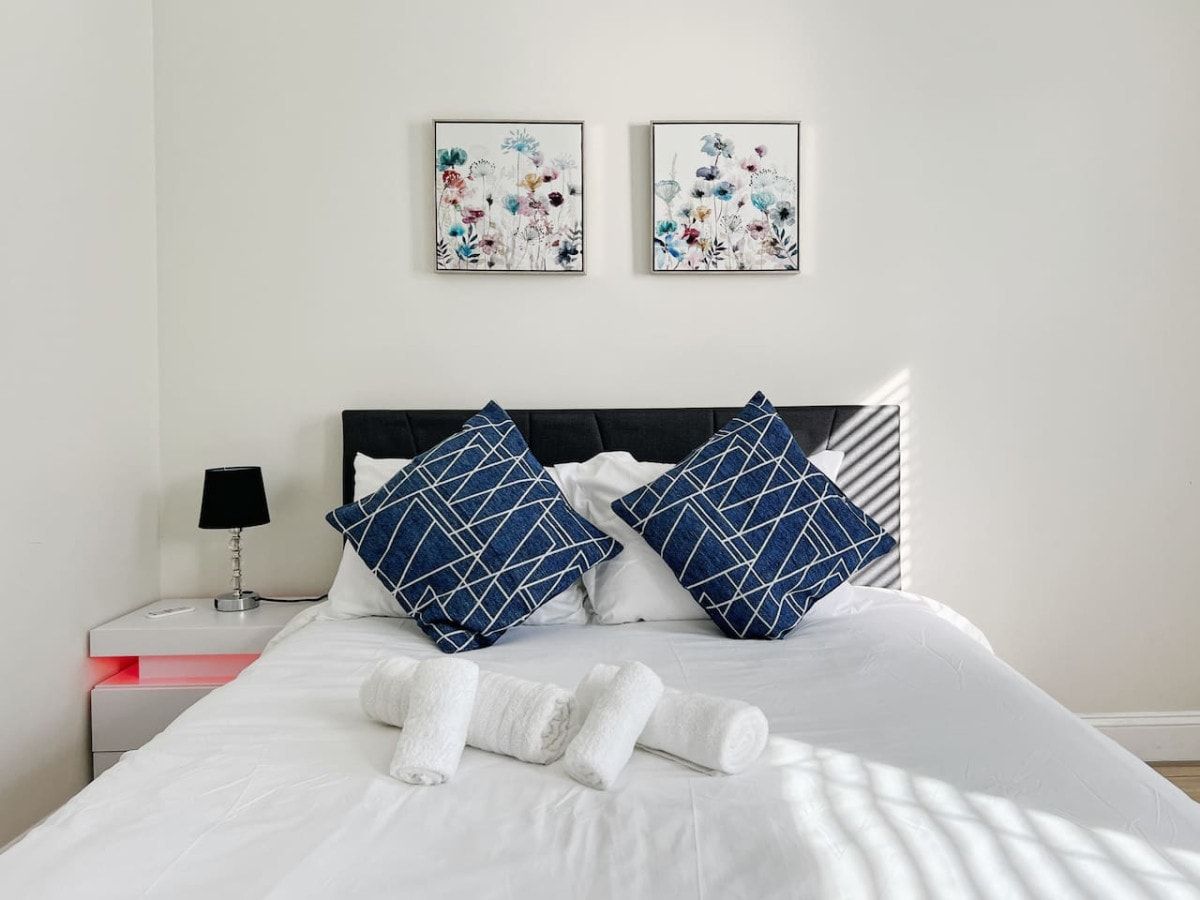 A neatly made bed with white linens and two pillows, a bedside table with a lamp, and two framed floral artworks on the wall.