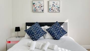 A neatly made bed with white linens and two pillows, a bedside table with a lamp, and two framed floral artworks on the wall.