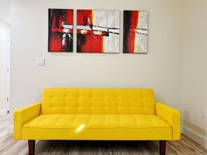 A bright yellow sofa in a room with a white wall and three abstract paintings above it. - Ramblei near Drexel Park: 4-Bedroom Unit A (Philadelphia)
