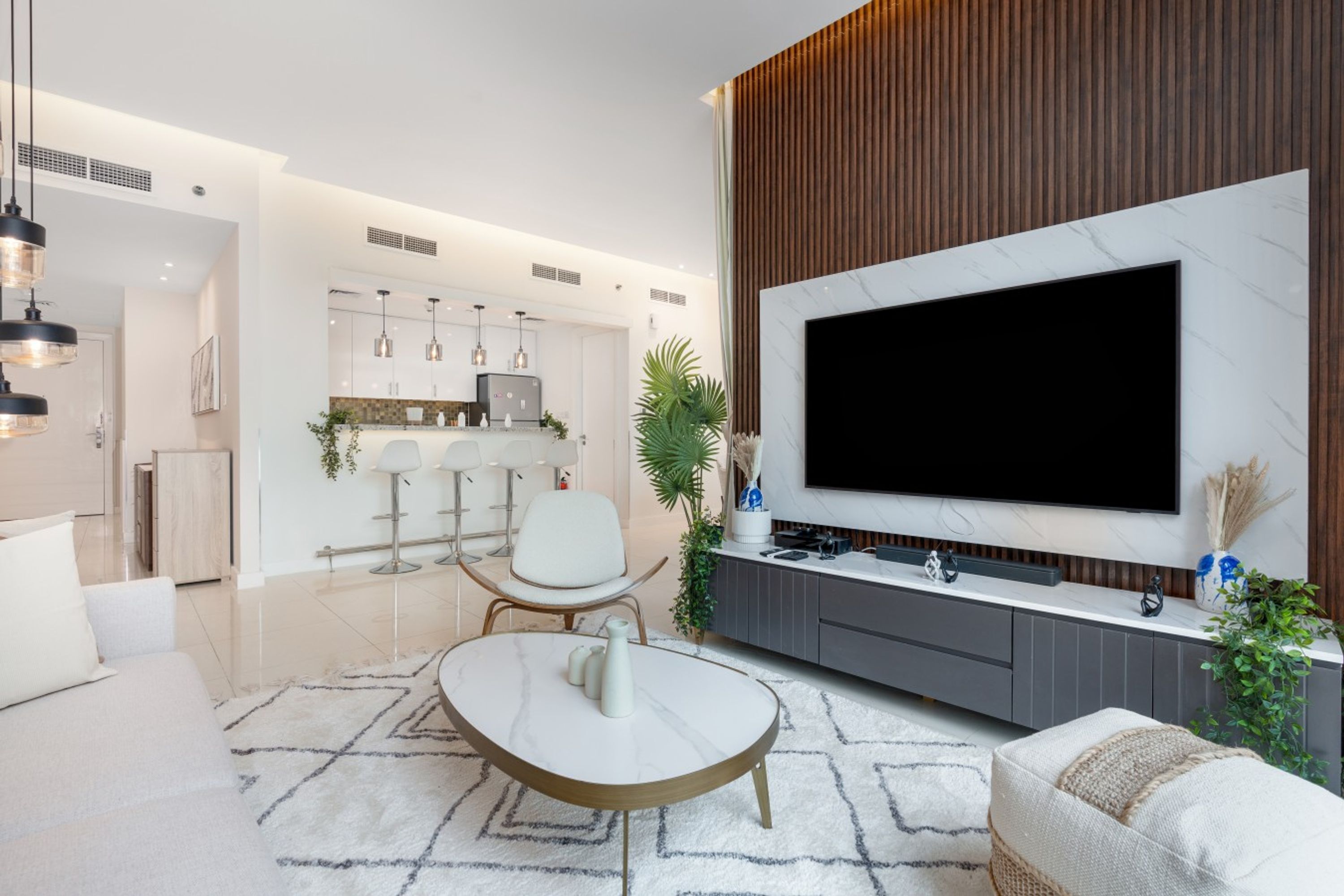 A modern living room with a large flat-screen TV, a dining area with bar stools, and a kitchen with a built-in fridge.