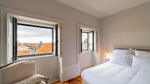 Une chambre à coucher comprenant un lit, une chaise, une table de chevet et deux fenêtres donnant sur la ville.
