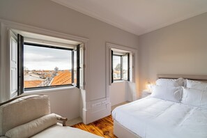 Ein Schlafzimmer mit einem Bett, einem Sessel, einem Nachttisch und zwei Fenstern mit Blick auf die Stadt.