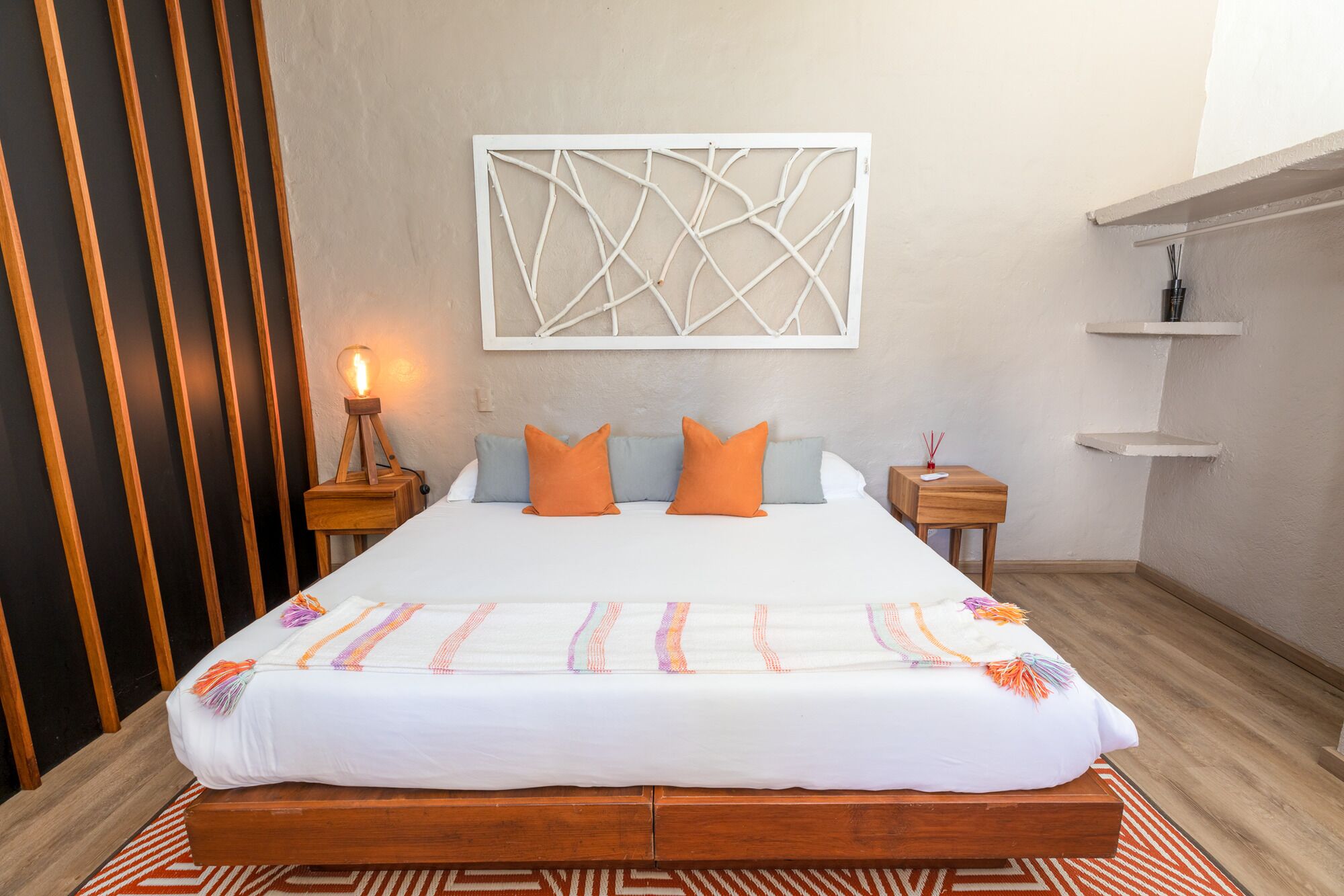 A bed with a white mattress, orange and blue pillows, a decorative wall piece, a bedside lamp, and a small wooden nightstand.