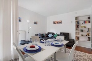 Een eethoek met een witte tafel gedekt voor twee personen, een boekenkast en een grijze bank op de achtergrond.