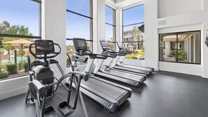 Een sportschool met loopbanden, elliptische trainers en een hometrainer, grote ramen en uitzicht op een zwembad en gebouwen.