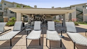 Een overdekt terras met lounge fauteuils en tafels, een stenen muur met geometrische patronen, en een trap aan de rechterkant.