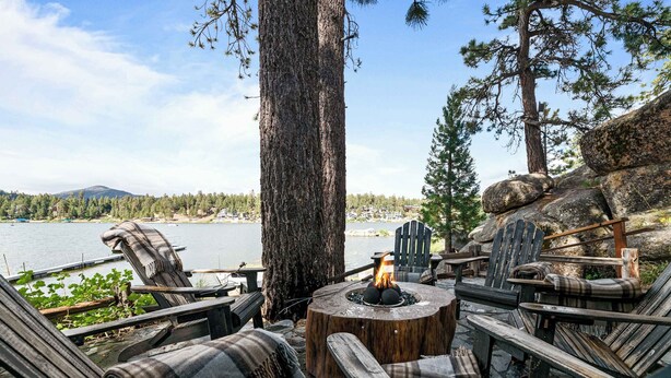 Een houten terras met Adirondack stoelen, een vuurkorf in het midden en uitzicht op een meer en bos.