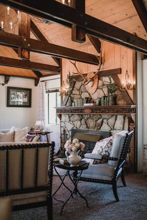 A cozy room with a fireplace, wooden beams, a stone wall, and a seating area with a table and chairs.