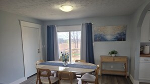 A dining area with a round table, four chairs, a vase with flowers, a painting on the wall, and a wooden console table with a plant.