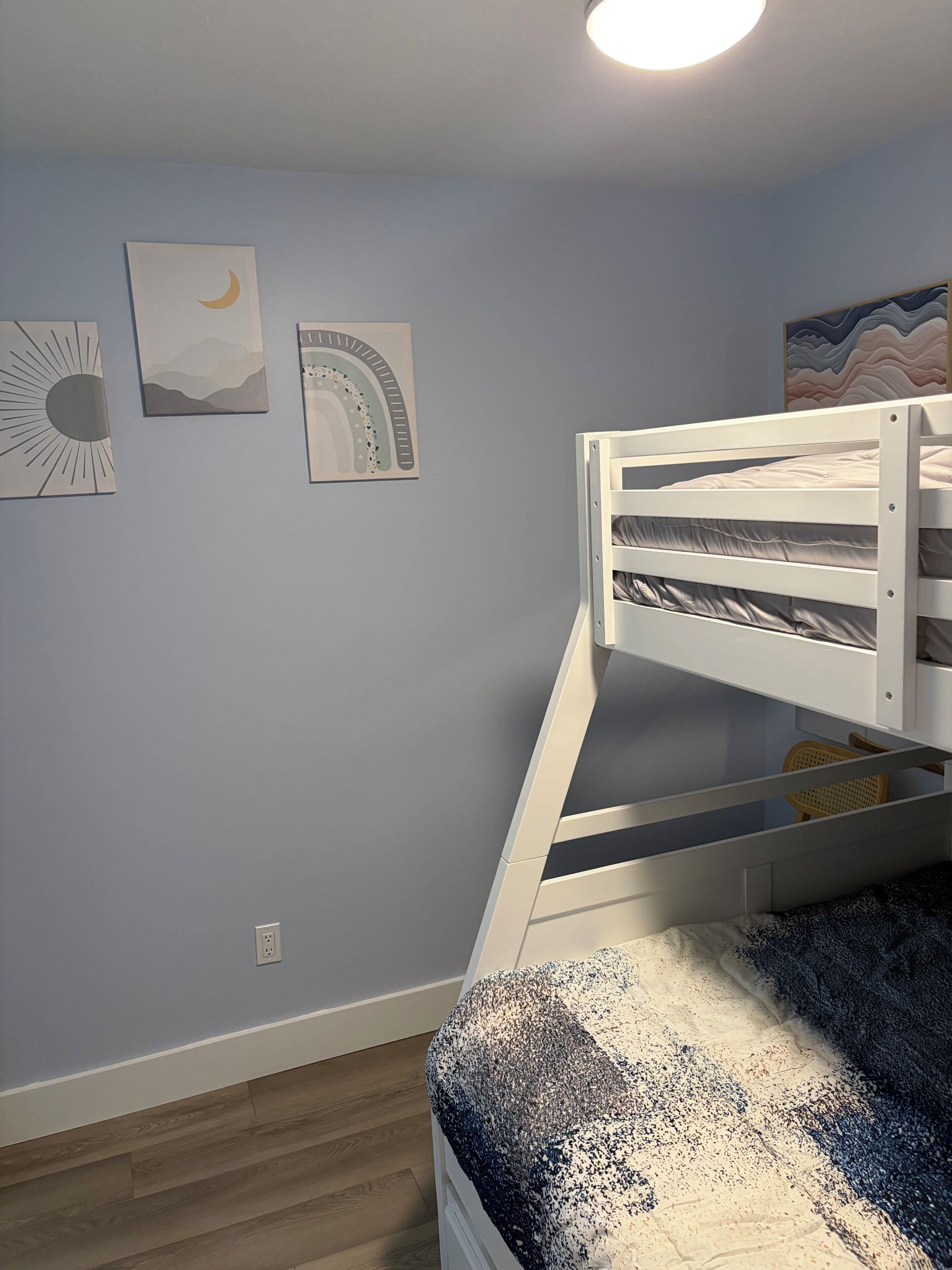 Una cama litera con estructura blanca, una pared con tres obras de arte enmarcadas, y un suelo de madera.
