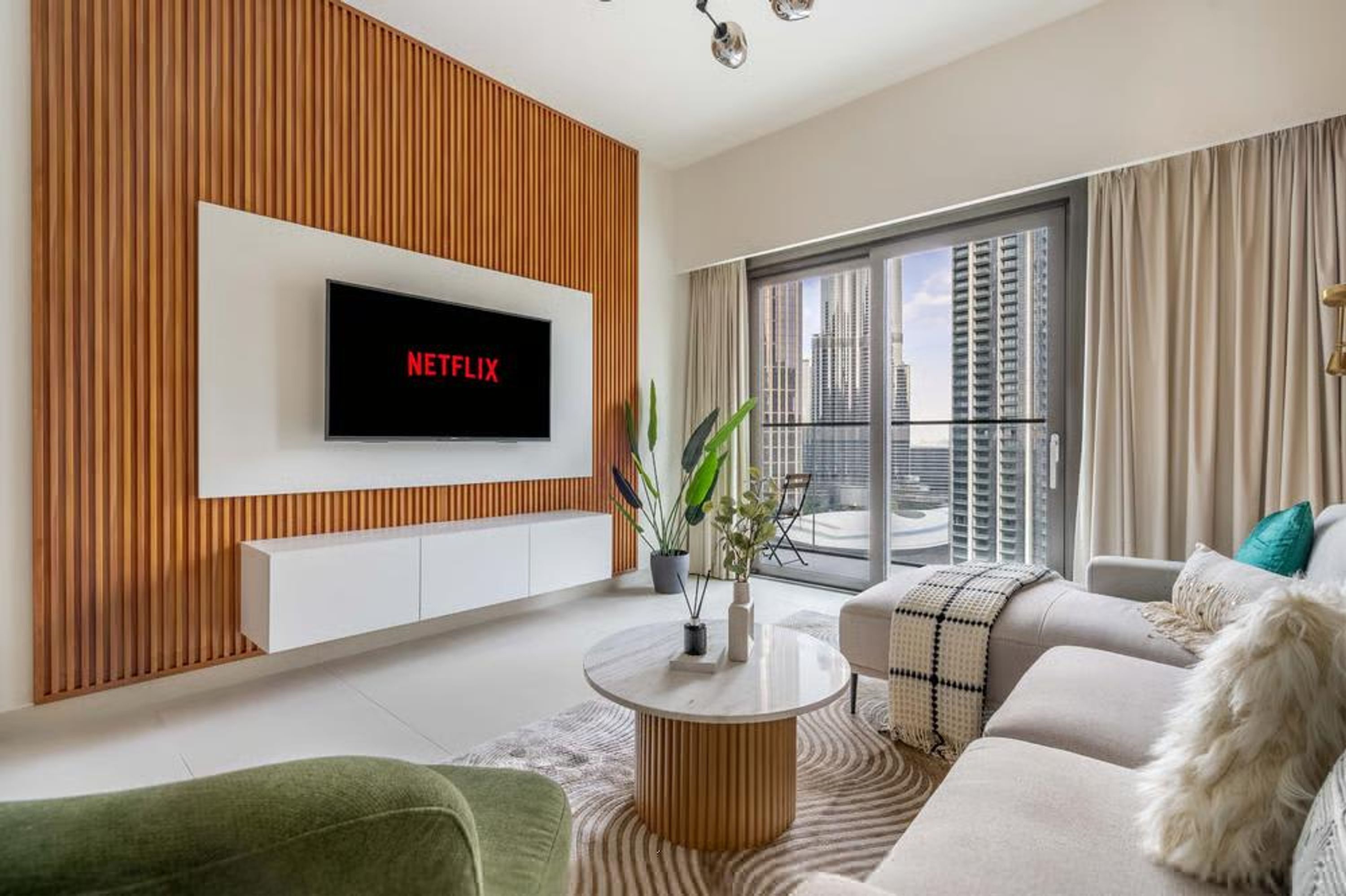 Una sala de estar moderna con una televisión montada que muestra 'NETFLIX', un sofá, una mesa de centro y vistas a un paisaje urbano a través de ventanas amplias.