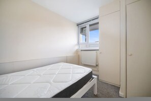 A small, clean room with a single bed, a wardrobe, and a window with a view of buildings. - 4-Bed Maisonette with Balcony (Greater London)