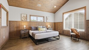 Un dormitorio con techo de madera, una cama con almohadas, una mesita de noche, una silla y una ventana con persianas.