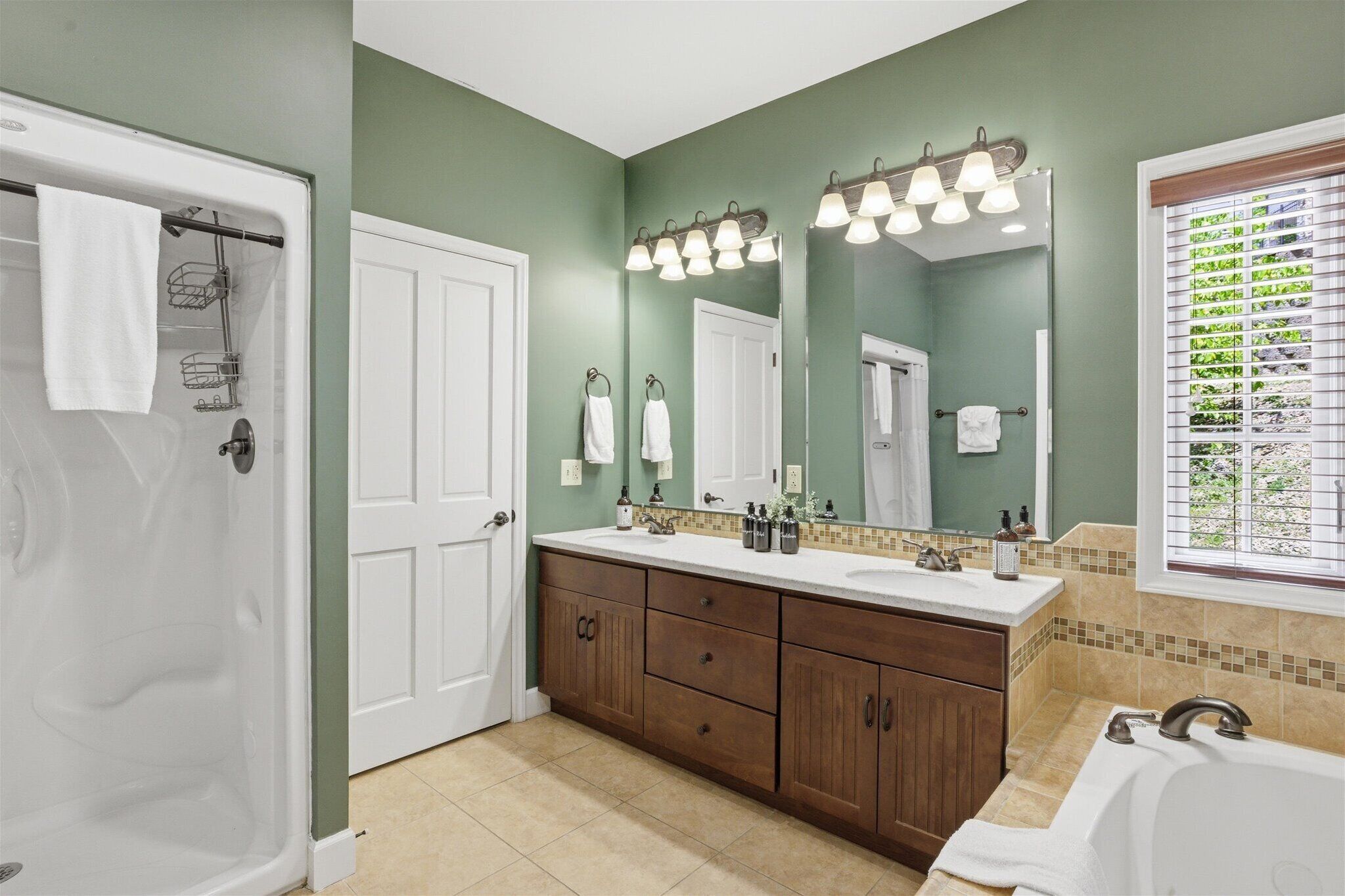 A bathroom with a shower, a double sink vanity, a large mirror, and a window with blinds.