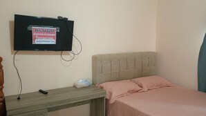 Een slaapkamer met een bed, een bureau met lades, een televisie aan de muur en een wandplank.
