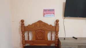 Een enkel bed met houten hoofdbord, een klein bureau met lades, een aan de muur bevestigde televisie, en een bord met advertenties voor tijdelijke appartementen.