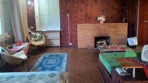 A cozy room with a fireplace, wooden walls, a wicker chair, a table with a tablecloth, and a blue patterned rug.