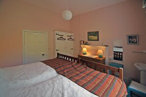 A bedroom with a bed, a desk with a lamp, a wardrobe, a mirror, and a shelf with books.