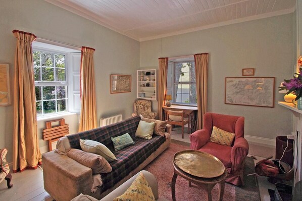 A cozy living room with a sofa, armchair, and a small table. There is a window with curtains, a radiator, and a bookshelf.