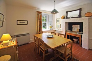 A dining area with a wooden table, chairs, a window with curtains, a radiator, a lamp, and a fireplace with a mirror above it.