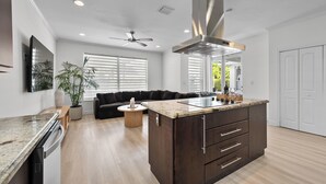 Eine moderne Küche mit einer zentralen Kochinsel, Holzschränken, einem Deckenventilator und einem Essbereich mit einer Couch und einem Couchtisch.