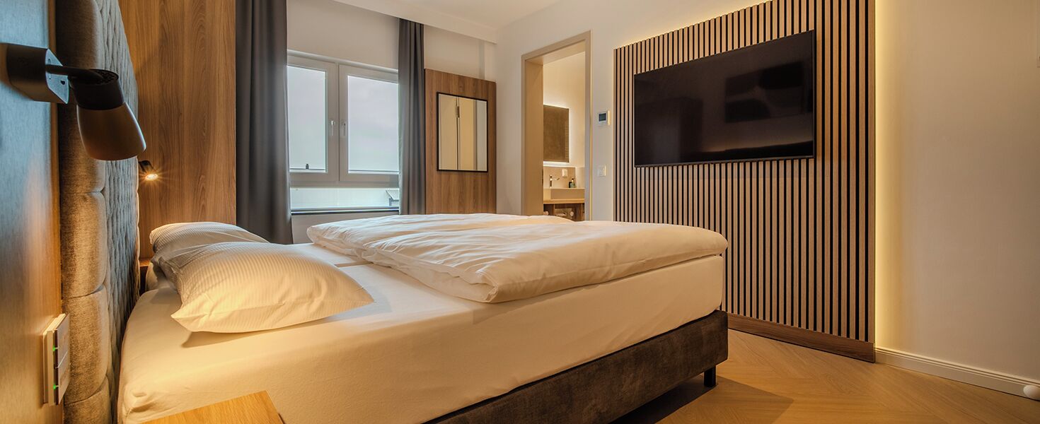 A modern hotel room with a large bed, a flat-screen TV mounted on the wall, and a wooden accent wall.
