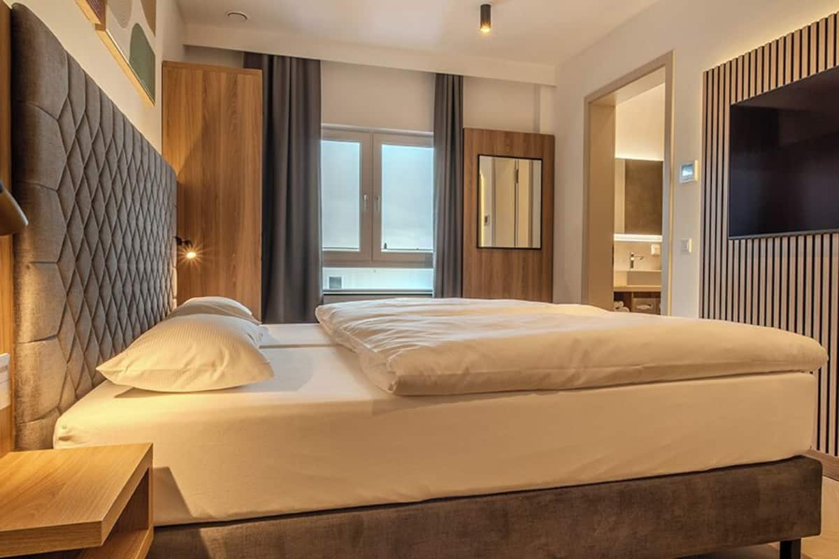 Una habitación de hotel moderna con una cama, un televisor, un armario de madera y un baño visible a través de una puerta abierta.