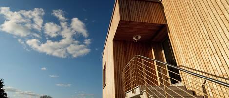 Un edificio moderno con fachada de madera, una escalera que lleva a un nivel superior y un cielo azul despejado con nubes.